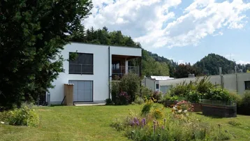 Modernes Haus mit weißer Fassade und gepflegtem Gartenbereich vor grüner Bergkulisse.