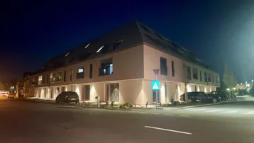 Modernes Gebäude bei Nacht mit beleuchteter Fassade und Mansarddach an einer Straßenecke.