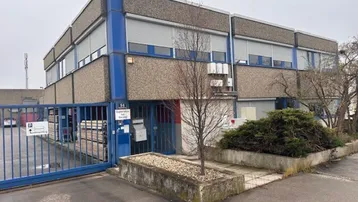 Außenansicht eines zweistöckigen Gewerbegebäudes mit Betonfassade, blauen Akzenten und Rollläden. Ein umzäunter Eingangsbereich ist sichtbar.