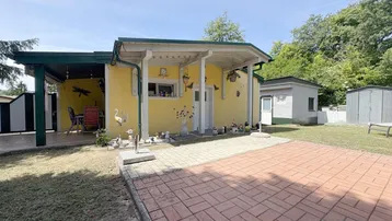 Gelbes Haus mit überdachtem Eingangsbereich, gepflastertem Vorplatz und Rasenfläche unter blauem Himmel.