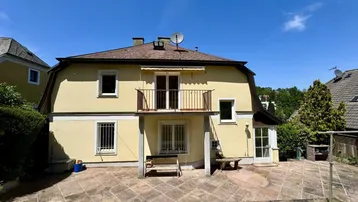 Rückseite des Hauses mit großer gepflasterter Terrasse und Balkon im Obergeschoss unter blauem Himmel.