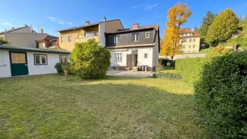Großer Gartenbereich mit Rasenfläche und kleiner Terrasse hinter dem Haus unter blauem Himmel.