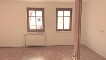 Heller Wohnraum mit zwei Holzfenstern, Parkettboden und Heizkörper, bietet viel Tageslicht.