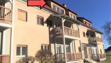 Mehrfamilienhaus mit mehreren Balkonen und Dachgauben unter blauem Himmel und grüner Umgebung.