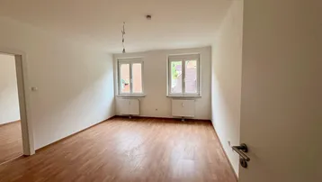 Helles, leeres Zimmer mit Laminatboden und zwei Fenstern, bereit zur individuellen Gestaltung.
