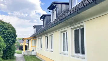 Seitenansicht des Gebäudes mit hellgelber Fassade, Dachgauben und einem gepflegten Gartenweg unter blauem Himmel.
