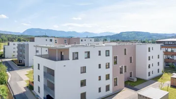 Moderne Wohngebäude in einer Siedlung mit Bergblick unter blauem Himmel und grüner Umgebung.