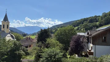 Weitläufiger Ausblick von einem Balkon auf eine Kirche, Häuser und die umliegende Berglandschaft.