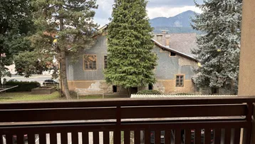 Blick vom Balkon auf ein älteres Gebäude mit Bäumen und Bergen im Hintergrund unter bewölktem Himmel.