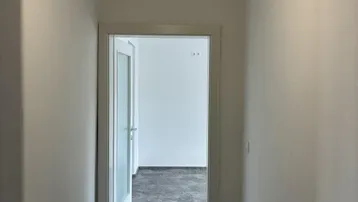 Schmaler Flur mit dunklem Boden und weiß gestrichenen Wänden, der zu einem weiteren Raum führt.