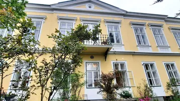 Historisches Gebäude mit gelber Fassade, weißen Fensterrahmen und üppiger Begrünung.