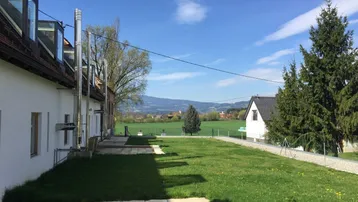 Weitläufiger Garten mit Rasenfläche und Terrasse, umgeben von grünen Feldern und Bergen.