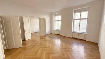 Helles Zimmer mit Fischgrätparkettboden und hohen Fenstern, das viel Tageslicht hereinlässt.