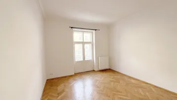 Helles Zimmer mit Fischgrätparkett, hohen Decken und Stuckelementen an der Decke.
