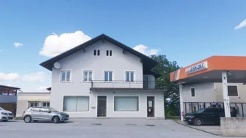 Gebäude mit weißer Fassade, Satteldach und Balkon, umgeben von Parkplätzen und einer Tankstelle.