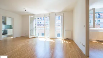 Helles, leeres Wohnzimmer mit Parkettboden und großen Fenstern, die viel Tageslicht hereinlassen.