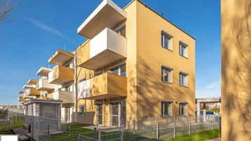 Modernes Mehrfamilienhaus mit gelber Fassade, Balkonen und gepflegter Grünfläche unter blauem Himmel.