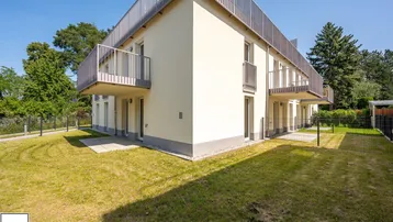 Modernes Mehrfamilienhaus mit hellbeiger Fassade, Balkonen und umliegender Grünfläche unter blauem Himmel.