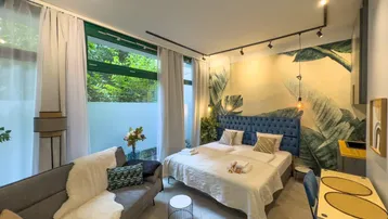 Modernes Schlafzimmer mit Doppelbett, Sofa und tropischer Wandtapete, hell durch große Fenster.