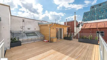 Große Dachterrasse mit Holzbelag, Pflanzenkübeln und Blick auf die Stadt unter blauem Himmel.