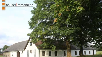 Ländliches Anwesen mit traditioneller Architektur und großen Bäumen im Vordergrund.