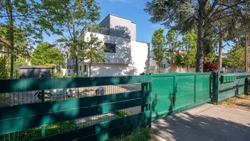 Modernes Wohngebäude mit weißer Fassade und Flachdach, umgeben von Bäumen und einem grünen Zaun.