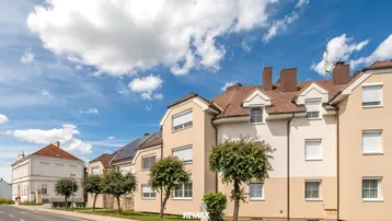 Mehrfamilienhaus mit beiger Fassade, Rollläden und Solaranlagen unter blauem Himmel.
