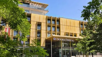 Moderne Hotelfassade mit Holzverkleidung und großen Fenstern, umgeben von grünen Bäumen unter blauem Himmel.