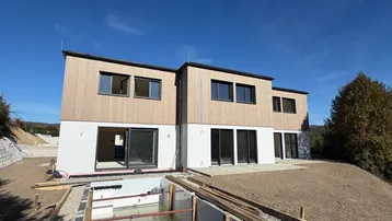 Moderne Neubauimmobilie mit Holzfassade und weißem Putz, umgeben von unfertiger Außenanlage und Baugrube.