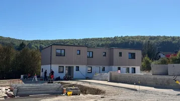 Neubau in ländlicher Umgebung mit Wald im Hintergrund und Baustelle im Vordergrund.