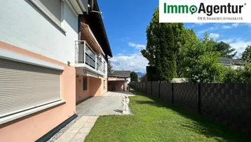 Seitenansicht des Hauses mit gepflegtem Rasen, gepflastertem Bereich und Balkon im Obergeschoss.