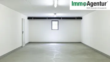 Leerer Garagenraum mit weißen Wänden, Betonboden, einem kleinen Fenster und Deckenbeleuchtung.