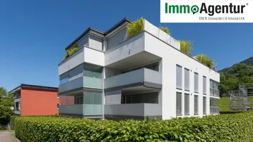 Modernes Mehrfamilienhaus mit weißer Fassade, großen Fenstern und begrüntem Flachdach in sonniger Umgebung.