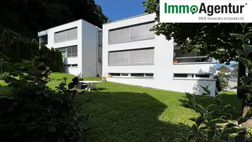 Modernes weißes Mehrfamilienhaus mit großen Fenstern und grüner Umgebung, eingebettet in die Natur.