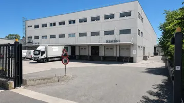 Großes Industriegebäude mit mehreren Laderampen und einem Lastwagen auf dem Vorplatz unter blauem Himmel.