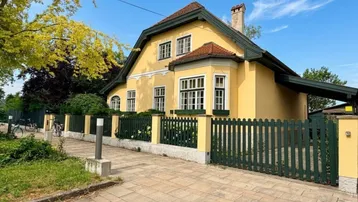 Gelbe Villa mit grünem Zaun und gepflastertem Gehweg unter blauem Himmel.