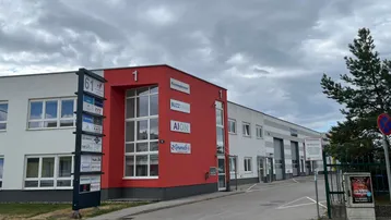 Gewerbegebäude mit markanter rot-weißer Fassade, großen Fenstern und Firmenbeschilderung an einem bewölkten Tag.
