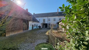 Alter Bauernhof mit gepflastertem Innenhof und traditioneller Architektur unter strahlendem Sonnenschein.