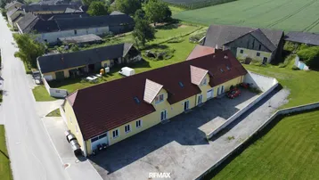 Luftaufnahme eines langen, gelben Gebäudes mit rotem Ziegeldach, umgeben von ländlicher Landschaft und anderen Gebäuden.