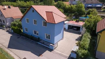 Luftaufnahme eines blauen Einfamilienhauses mit rotem Dach und gepflasterter Einfahrt.