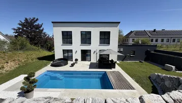 Modernes Einfamilienhaus mit Swimmingpool, Holzdeck und weitläufigem Garten unter strahlend blauem Himmel.