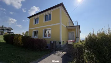 Gelbes Einfamilienhaus mit Balkon und gepflegtem Vorgarten unter blauem Himmel.