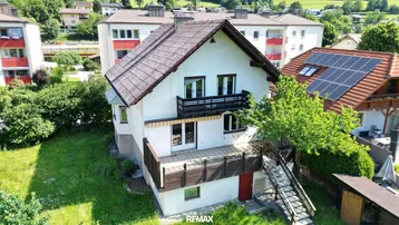 Freistehendes Haus mit Balkonen und Terrasse, umgeben von Grünflächen und Nachbargebäuden.