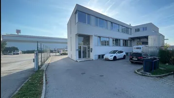 Modernes weißes Bürogebäude mit großen Fenstern und einem geräumigen Parkplatz unter blauem Himmel.