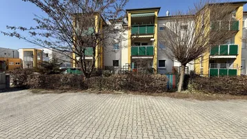 Mehrfamilienhaus mit gelber Fassade, grünen Balkonen und gepflastertem Vorplatz.