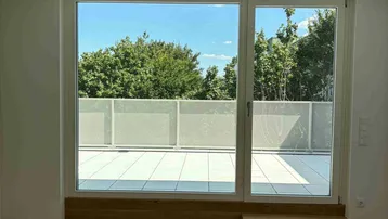 Helles Zimmer mit Holzboden und Zugang zu einer großen Terrasse mit Blick ins Grüne.
