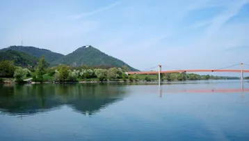 Malerischer Blick auf einen Fluss mit Brücke und bewaldeten Hügeln im Hintergrund unter blauem Himmel.