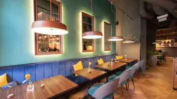 Gemütlicher Restaurantbereich mit blauer Samtbank, Holztischen und grünen Wänden, beleuchtet von modernen Pendelleuchten.