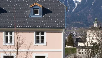 Nahaufnahme der Hausfassade mit Dachgaube und beeindruckendem Alpenpanorama im Hintergrund.