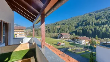 Balkon mit Holzdach und weitem Blick auf grüne Berge, Wald und umliegende Häuser.